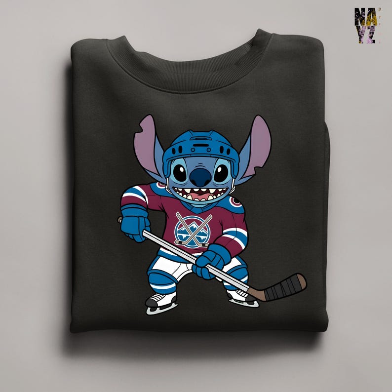 Puede incluir: Sudadera negra con un personaje de dibujos animados con uniforme de hockey. El personaje azul con orejas grandes lleva un casco. La camiseta de hockey es granate, azul y blanca, con un logotipo del equipo. Sostiene un palo de hockey.