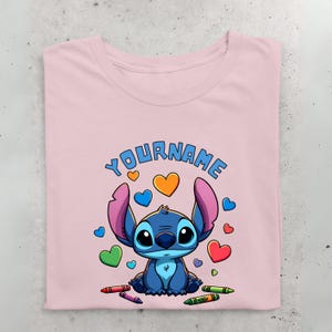 K&ouml;nnte beinhalten: Rosa T-Shirt mit einer blauen Stitch-Figur aus Lilo & Stitch. Stitch ist von bunten Herzen und Buntstiften umgeben. Der Text "YOUR NAME" befindet sich &uuml;ber Stitch.