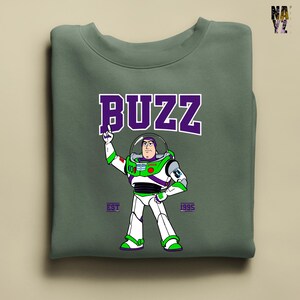 May include: Sage green sweatshirt featuring a graphic of Buzz Lightyear in his spacesuit, with the word "BUZZ" in purple block letters above. The text "EST 1995" is below the character. The sweatshirt is folded.