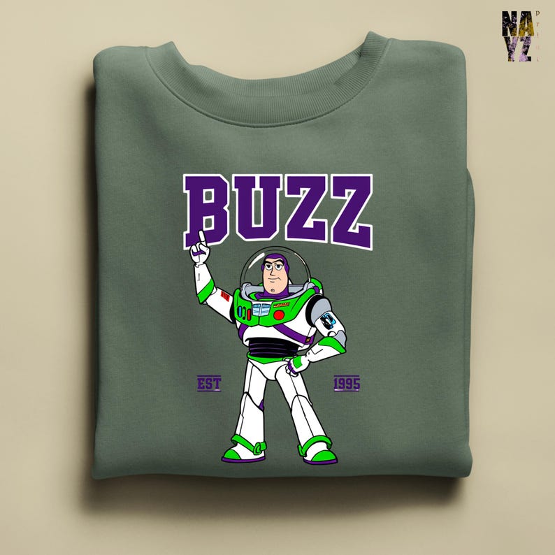 May include: Sage green sweatshirt featuring a graphic of Buzz Lightyear in his spacesuit, with the word "BUZZ" in purple block letters above. The text "EST 1995" is below the character. The sweatshirt is folded.