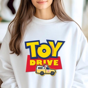 May include: White sweatshirt with a blue and yellow "Toy Drive" graphic featuring a cartoon toy truck.