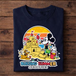 May include: Navy blue t-shirt featuring a colorful graphic of Mickey Mouse building a sandcastle with Pluto. The design includes a sunset backdrop and the text "Building Summer Memories".