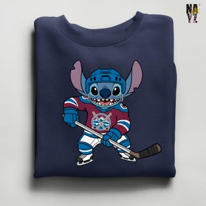 Puede incluir: Sudadera azul marino con un personaje de dibujos animados con uniforme de hockey. El personaje es azul con orejas grandes y sostiene un palo de hockey. El uniforme es burdeos, azul y blanco, con un logotipo del equipo en el pecho.