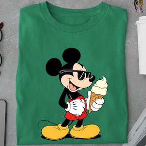 Puede incluir: Camiseta verde con un dise&ntilde;o de dibujos animados de Mickey Mouse con gafas de sol y un helado. Mickey est&aacute; representado en negro, rojo, amarillo y blanco. La camiseta est&aacute; doblada sobre una superficie gris.