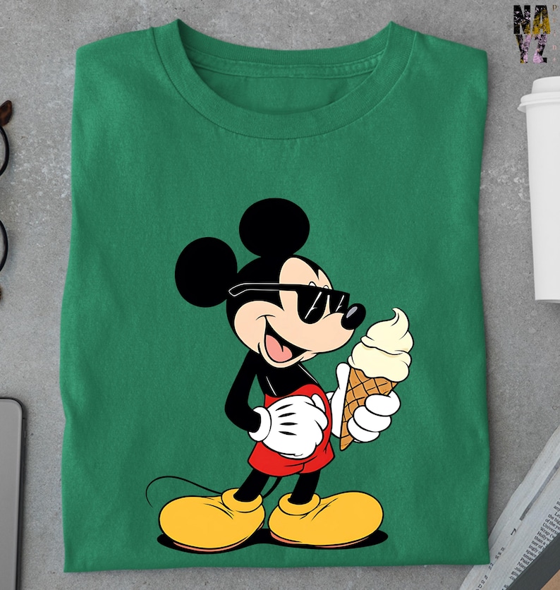 Puede incluir: Camiseta verde con un dise&ntilde;o de dibujos animados de Mickey Mouse con gafas de sol y un helado. Mickey est&aacute; representado en negro, rojo, amarillo y blanco. La camiseta est&aacute; doblada sobre una superficie gris.