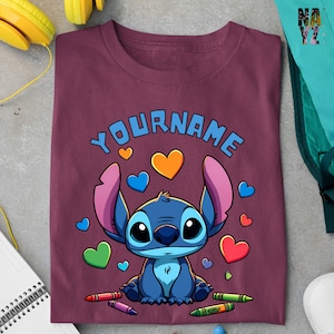 K&ouml;nnte beinhalten: Ein maroonfarbenes T-Shirt mit einer blauen Comicfigur mit gro&szlig;en Augen und rosa Ohren. Die Figur ist von bunten Herzen und Buntstiften umgeben. Der Text "YOURNAME" ist &uuml;ber der Figur gedruckt.
