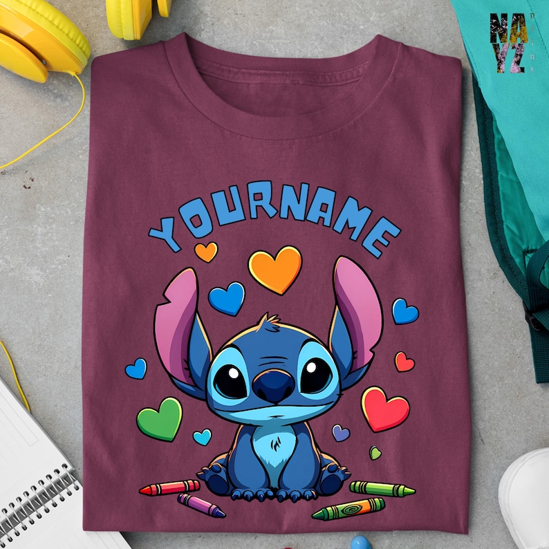 K&ouml;nnte beinhalten: Ein maroonfarbenes T-Shirt mit einer blauen Comicfigur mit gro&szlig;en Augen und rosa Ohren. Die Figur ist von bunten Herzen und Buntstiften umgeben. Der Text "YOURNAME" ist &uuml;ber der Figur gedruckt.
