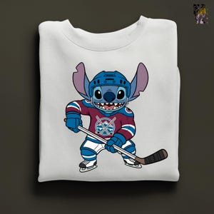 Puede incluir: Sudadera blanca con un personaje de dibujos animados con uniforme de hockey. El personaje azul con orejas grandes lleva casco y camiseta con el logo del equipo. El personaje sostiene un palo de hockey y lleva patines.