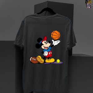 May include: Dark gray t-shirt featuring a cartoon Mickey Mouse character in a sports theme. Mickey is wearing a red hat, blue shirt, and red shorts, holding a basketball. A football, tennis racket, and tennis ball are also visible.