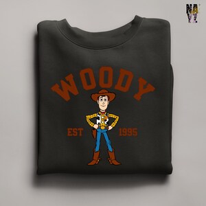 Puede incluir: Sudadera negra con una imagen de dibujos animados de Woody de Toy Story. La palabra "WOODY" est&aacute; arqueada en la parte superior en letras marrones, con "EST 1995" debajo. La sudadera est&aacute; doblada.