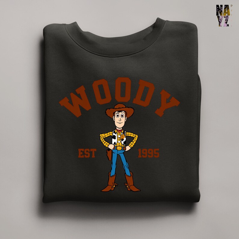 Puede incluir: Sudadera negra con una imagen de dibujos animados de Woody de Toy Story. La palabra "WOODY" est&aacute; arqueada en la parte superior en letras marrones, con "EST 1995" debajo. La sudadera est&aacute; doblada.