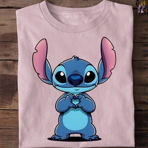 May include: A light pink t-shirt featuring a cartoon character, Stitch, in shades of blue. Stitch is depicted with large black eyes, a blue nose, and large ears. His hands are forming a heart shape. The shirt is folded on a wooden surface.