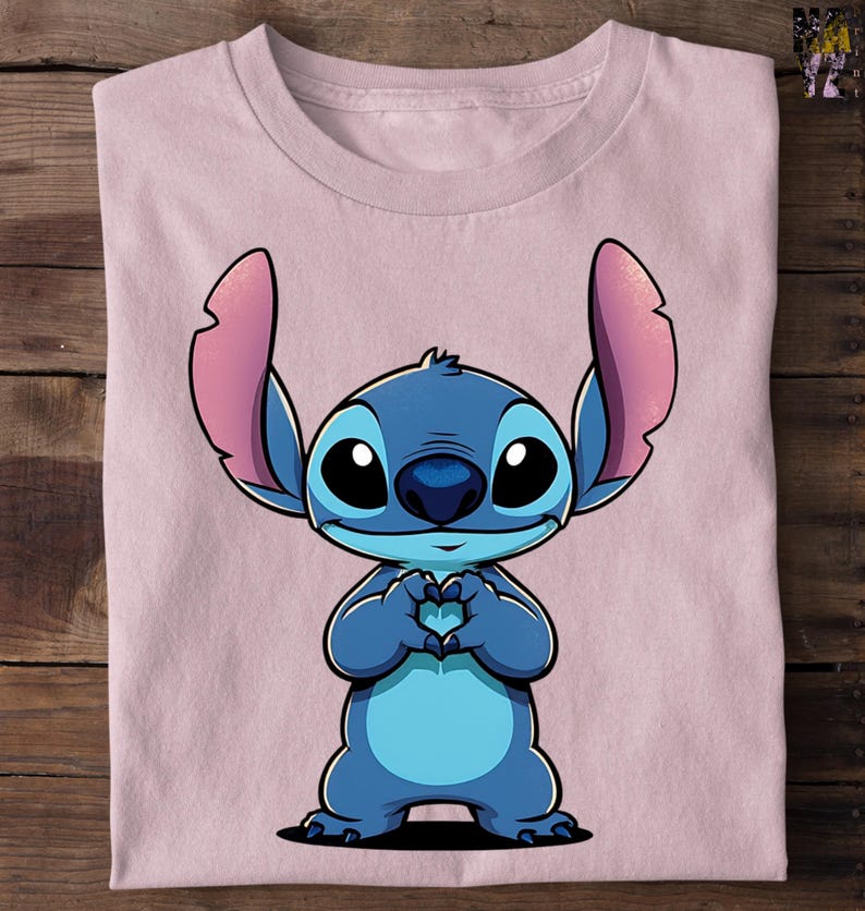 May include: A light pink t-shirt featuring a cartoon character, Stitch, in shades of blue. Stitch is depicted with large black eyes, a blue nose, and large ears. His hands are forming a heart shape. The shirt is folded on a wooden surface.