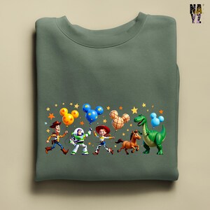 Puede incluir: Sudadera verde salvia con un gr&aacute;fico colorido de personajes de Toy Story sosteniendo globos con forma de Mickey Mouse. Woody, Buzz Lightyear, Jessie, un caballo y un dinosaurio est&aacute;n representados con estrellas.