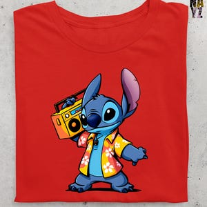 Op de afbeelding: Rood T-shirt met een cartoon illustratie van Stitch uit Lilo & Stitch. Stitch is blauw, draagt een geel Hawaiiaans shirt met witte bloemen en houdt een gele boombox vast. Het shirt is opgevouwen en heeft een eenvoudig ontwerp.