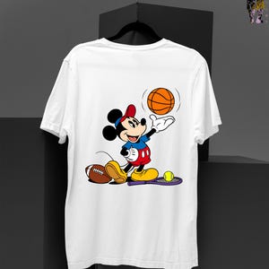 May include: White t-shirt featuring a cartoon Mickey Mouse character in a red hat and blue shirt, playing with a basketball. A football and tennis racket with a ball are also present. The design is colorful and playful.