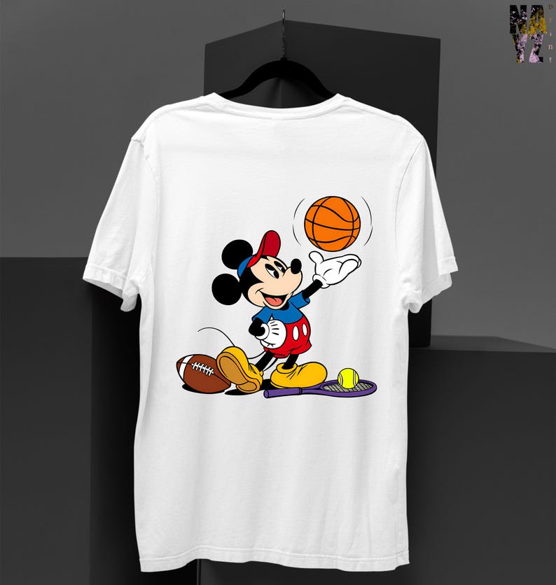 May include: White t-shirt featuring a cartoon Mickey Mouse character in a red hat and blue shirt, playing with a basketball. A football and tennis racket with a ball are also present. The design is colorful and playful.