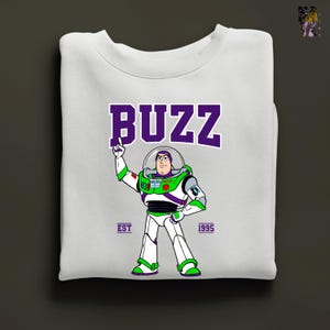May include: White sweatshirt featuring a graphic of Buzz Lightyear in his spacesuit, with the word "BUZZ" in purple block letters above. The design includes the text "EST 1995". The sweatshirt is folded neatly.
