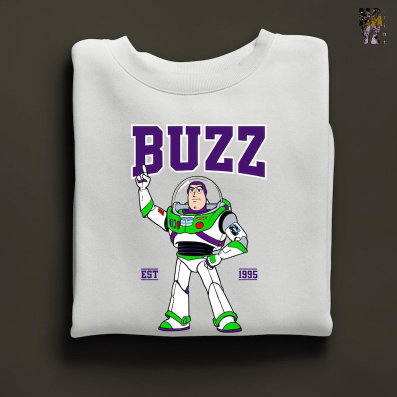 May include: White sweatshirt featuring a graphic of Buzz Lightyear in his spacesuit, with the word "BUZZ" in purple block letters above. The design includes the text "EST 1995". The sweatshirt is folded neatly.