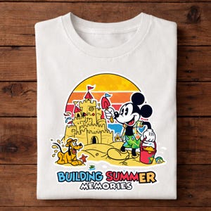 May include: White t-shirt featuring a colorful graphic of Mickey Mouse building a sandcastle with Pluto at the beach. The design includes a sunset backdrop and the text "BUILDING SUMMER MEMORIES".
