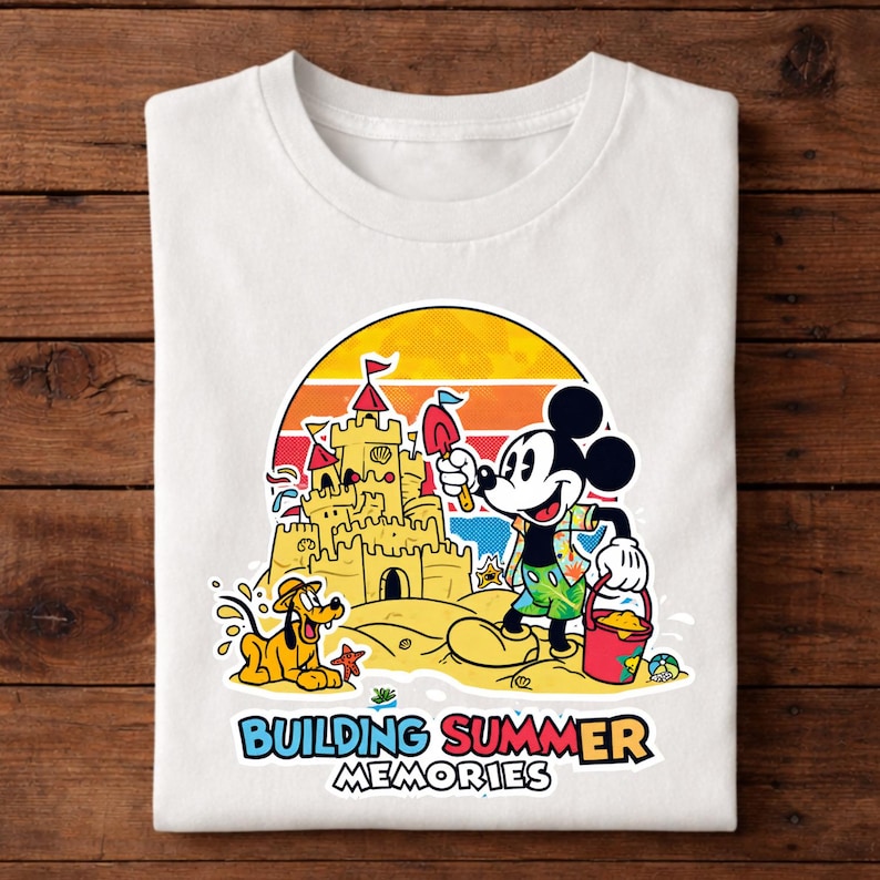 May include: White t-shirt featuring a colorful graphic of Mickey Mouse building a sandcastle with Pluto at the beach. The design includes a sunset backdrop and the text "BUILDING SUMMER MEMORIES".