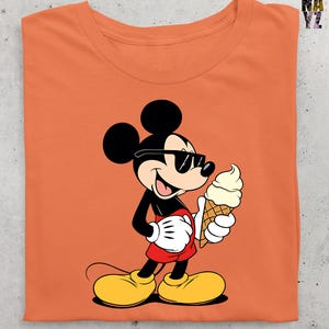 Puede incluir: Camiseta naranja con un dise&ntilde;o de dibujos animados de Mickey Mouse con gafas de sol y un helado. Mickey lleva pantalones cortos rojos, zapatos amarillos y guantes blancos.