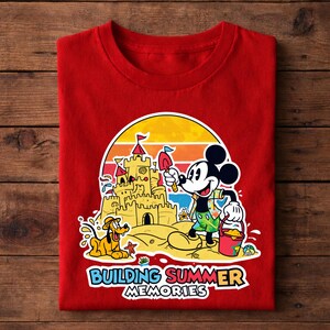May include: Red t-shirt featuring a colorful graphic of Mickey Mouse building a sandcastle with Pluto. The design includes a sunset backdrop and the text "BUILDING SUMMER MEMORIES".