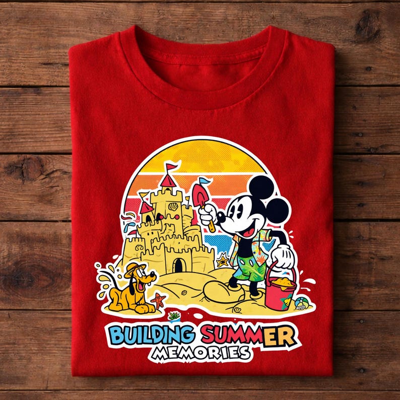 May include: Red t-shirt featuring a colorful graphic of Mickey Mouse building a sandcastle with Pluto. The design includes a sunset backdrop and the text "BUILDING SUMMER MEMORIES".