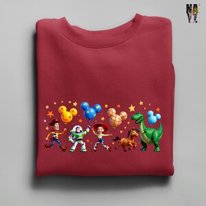 Puede incluir: Una sudadera granate con un gr&aacute;fico colorido de personajes de la franquicia Toy Story. El dise&ntilde;o incluye a Woody, Buzz Lightyear, Jessie, un dinosaurio y un caballo, junto con globos con forma de Mickey Mouse y estrellas.