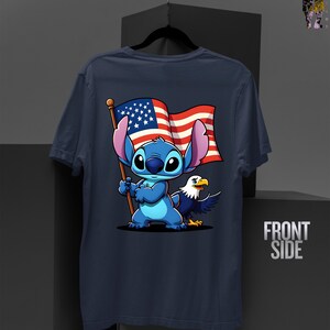 May include: Navy blue t-shirt featuring a cartoon character holding an American flag with an eagle. The character is blue with large ears. The flag has red and white stripes and a blue field with white stars. The eagle is white, blue, and yellow.