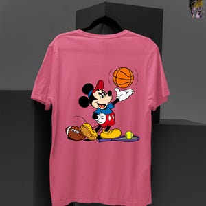 May include: Pink t-shirt featuring a cartoon Mickey Mouse character in a sports theme. Mickey is wearing a red cap, blue shirt, and red shorts, holding a basketball. A football, tennis racket, and tennis ball are also present.