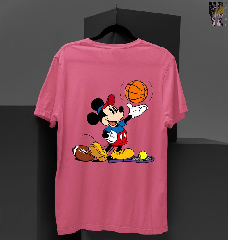 May include: Pink t-shirt featuring a cartoon Mickey Mouse character in a sports theme. Mickey is wearing a red cap, blue shirt, and red shorts, holding a basketball. A football, tennis racket, and tennis ball are also present.