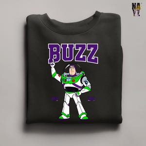 May include: Black sweatshirt featuring a cartoon illustration of Buzz Lightyear in a white and green spacesuit. The word "BUZZ" is printed above the character in purple block letters. The sweatshirt is a crew neck style.