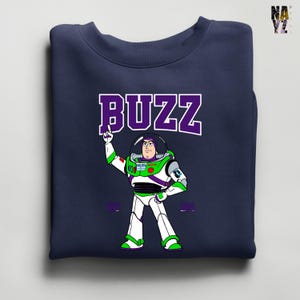 May include: Navy blue sweatshirt featuring a cartoon illustration of Buzz Lightyear from Toy Story. The word "BUZZ" is printed in large, purple letters above the character. Buzz is depicted in his signature white, green, and purple spacesuit, with his arm raised.