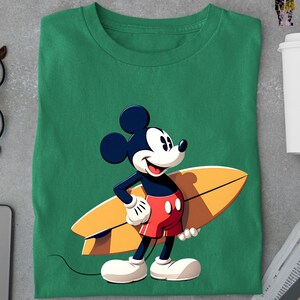 Op de afbeelding: Groen t-shirt met een cartoon illustratie van Mickey Mouse met een surfboard. Mickey draagt een rode broek, witte handschoenen en witte schoenen. Het surfboard is geel en bruin.