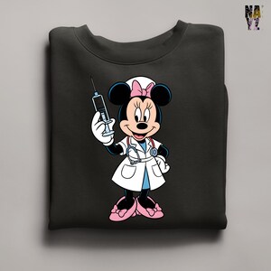 Peut inclure: Sweat-shirt noir avec un dessin anim&eacute; de Minnie Mouse habill&eacute;e en infirmi&egrave;re. Minnie Mouse porte une blouse blanche, des chaussures roses et un bonnet d'infirmi&egrave;re avec un n&oelig;ud rose. Elle tient une seringue. Le sweat-shirt est pli&eacute;.