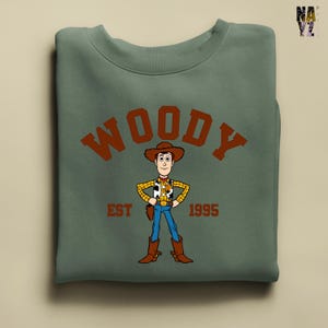 Puede incluir: Sudadera verde salvia con una ilustraci&oacute;n de dibujos animados de Woody de Toy Story. El dise&ntilde;o incluye la palabra "WOODY" en letras marrones arqueadas sobre el personaje, con "EST 1995" debajo. Woody est&aacute; representado con su atuendo de vaquero.