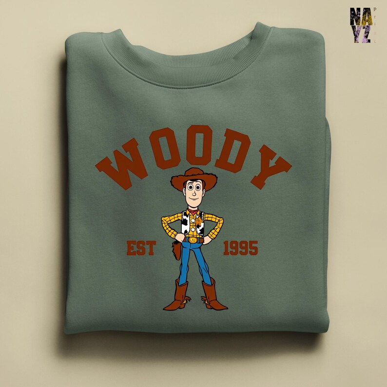 Puede incluir: Sudadera verde salvia con una ilustraci&oacute;n de dibujos animados de Woody de Toy Story. El dise&ntilde;o incluye la palabra "WOODY" en letras marrones arqueadas sobre el personaje, con "EST 1995" debajo. Woody est&aacute; representado con su atuendo de vaquero.