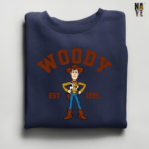 Puede incluir: Sudadera azul marino con una ilustraci&oacute;n de dibujos animados de Woody de Toy Story. El dise&ntilde;o incluye el nombre "WOODY" en letras marrones arqueadas, con "EST 1995" debajo. La sudadera est&aacute; doblada.