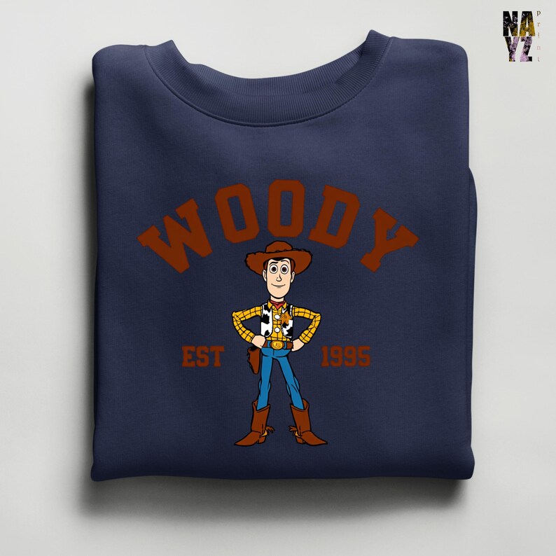Puede incluir: Sudadera azul marino con una ilustraci&oacute;n de dibujos animados de Woody de Toy Story. El dise&ntilde;o incluye el nombre "WOODY" en letras marrones arqueadas, con "EST 1995" debajo. La sudadera est&aacute; doblada.