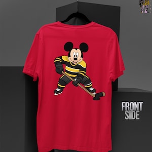 May include: Red t-shirt featuring a cartoon Mickey Mouse character in hockey gear, holding a hockey stick and puck. The character is wearing a black and yellow striped jersey and black shorts. The words "FRONT SIDE" are visible on the right.