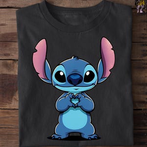 May include: Black t-shirt featuring a cartoon illustration of Stitch, a blue alien character with large ears and big eyes. Stitch is depicted with his hands forming a heart shape. The shirt is folded on a wooden surface.