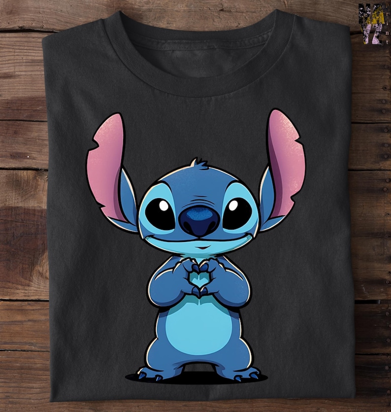 May include: Black t-shirt featuring a cartoon illustration of Stitch, a blue alien character with large ears and big eyes. Stitch is depicted with his hands forming a heart shape. The shirt is folded on a wooden surface.