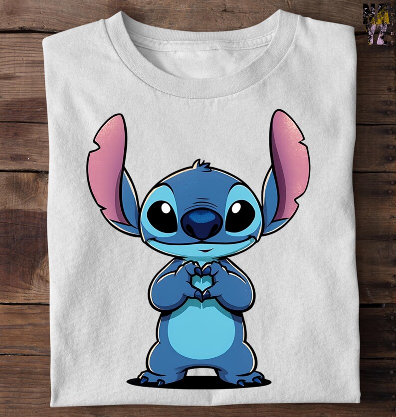 May include: White t-shirt featuring a cartoon character, Stitch, in shades of blue with pink ears. Stitch is making a heart shape with its hands. The shirt is folded on a wooden surface.