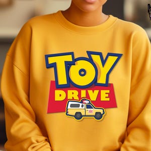 May include: Yellow sweatshirt with a Toy Story themed design. The design features the words "Toy Drive" in a blue and yellow font with a cartoon image of a yellow van with a red roof.