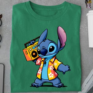 Op de afbeelding: Groen t-shirt met een cartoon illustratie van Stitch, die een gele boombox vasthoudt. Stitch draagt een Hawaiiaans shirt met een bloemenpatroon. Het ontwerp is gecentreerd op het shirt.