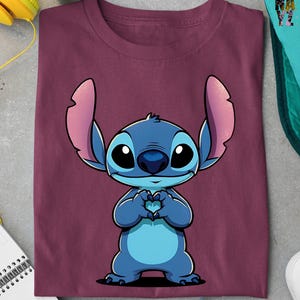May include: Burgundy t-shirt featuring a cartoon illustration of Stitch from Lilo & Stitch. The blue character is making a heart shape with its hands. The shirt is folded and has a soft appearance.