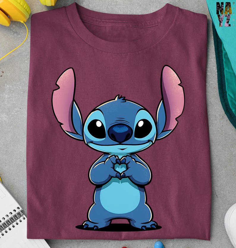 May include: Burgundy t-shirt featuring a cartoon illustration of Stitch from Lilo & Stitch. The blue character is making a heart shape with its hands. The shirt is folded and has a soft appearance.