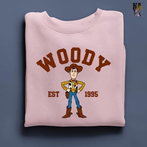 Puede incluir: Sudadera rosa claro con una ilustraci&oacute;n de dibujos animados de Woody de Toy Story. La palabra "WOODY" est&aacute; impresa en letras marrones sobre el personaje. Debajo est&aacute;n las palabras "EST 1995".