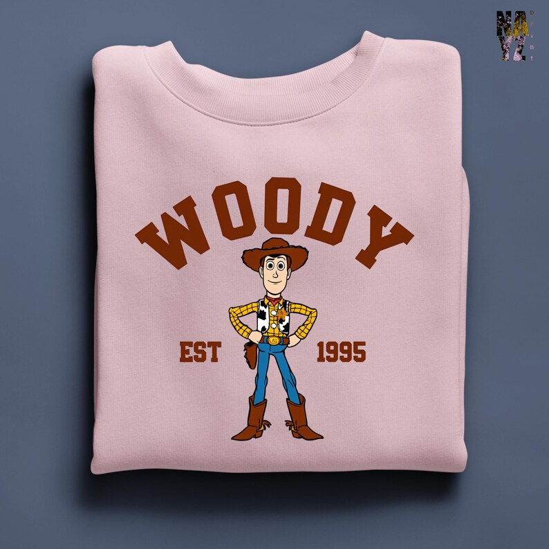 Puede incluir: Sudadera rosa claro con una ilustraci&oacute;n de dibujos animados de Woody de Toy Story. La palabra "WOODY" est&aacute; impresa en letras marrones sobre el personaje. Debajo est&aacute;n las palabras "EST 1995".